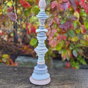 Op de afbeelding: Een verticale stapel gladde, ronde rivierstenen in verschillende tinten grijs, blauw en zachtroze. De stenen zijn op elkaar gebalanceerd en vormen een hoge, slanke toren die naar boven toe taps toeloopt en eindigt met een enkele kleine steen.