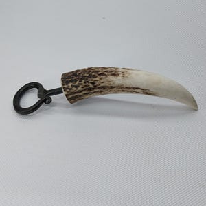 Pode incluir: Um abridor de garrafas com uma pega de chifre de veado castanho e branco. A pega tem a forma de um chifre de veado curvo. O abridor é feito de metal e tem um laço para abrir garrafas.