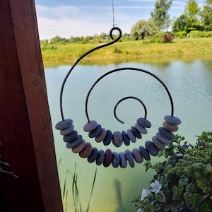 Pode incluir: Uma escultura decorativa suspensa com um design em espiral feito de metal escuro. A espiral é adornada com pedras de rio lisas cinzentas e castanhas. A escultura está pendurada em frente a um lago e vegetação.