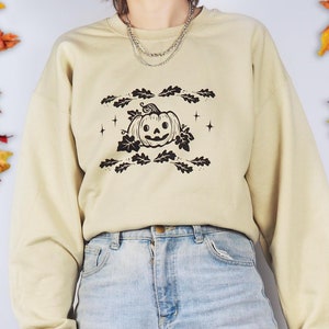 A photo of a woman wearing a beige jumper with an autumnal pumpkin surrounded by oak leaves printed on it.
