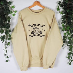 Pode incluir: Uma camisa de moletom bege com um gr&aacute;fico preto e branco de uma ab&oacute;bora com folhas e estrelas.