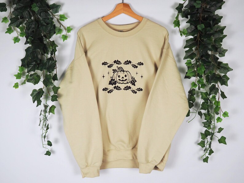 Pode incluir: Uma camisa de moletom bege com um gr&aacute;fico preto e branco de uma ab&oacute;bora com folhas e estrelas.