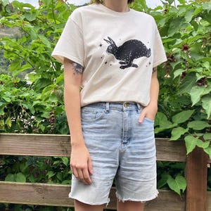 May include: A white t-shirt with a black rabbit graphic and stars. The rabbit is running and has a long tail. The shirt is being worn by a person with blue denim shorts.