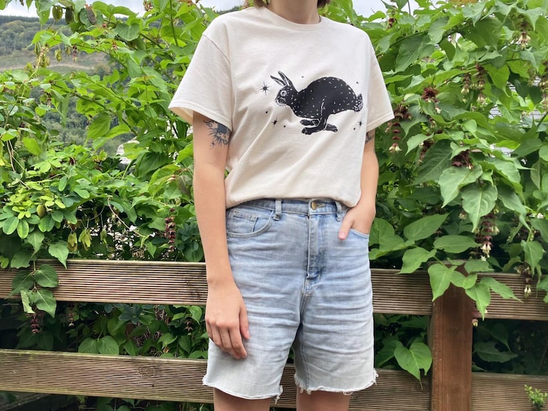 May include: A white t-shirt with a black rabbit graphic and stars. The rabbit is running and has a long tail. The shirt is being worn by a person with blue denim shorts.
