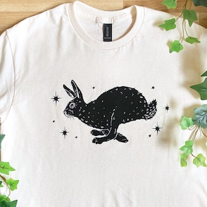 May include: A white t-shirt with a black silhouette of a rabbit running with stars printed on it.