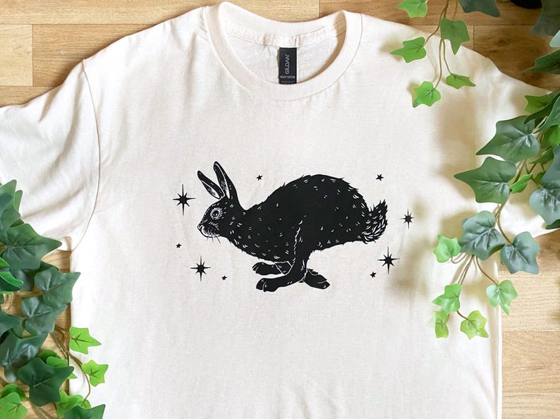 May include: A white t-shirt with a black silhouette of a rabbit running with stars printed on it.