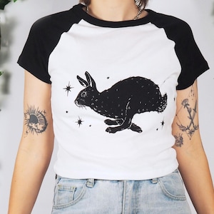 Könnte beinhalten: Ein weißes Raglan-T-Shirt mit schwarzen kurzen Ärmeln und einem schwarzen Rundhalsausschnitt. In der Mitte des T-Shirts befindet sich eine schwarze grafische Illustration eines laufenden Kaninchens, das von kleinen Sternen umgeben ist.