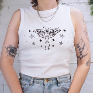 May include: White vest top featuring a black moth and star design. The moth is the central image, with detailed wing patterns and elongated lower sections. Stars of different shapes surround the moth.
