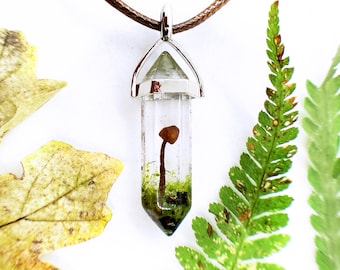 May include: A clear, crystal pendant with a small mushroom encased inside, suspended from a brown cord. The mushroom is brown with a long stem, surrounded by green moss. The pendant is accented with silver-colored metal. Dried leaves and ferns surround the pendant.