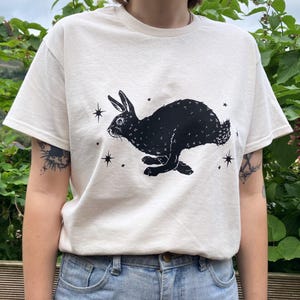 May include: A white t-shirt with a black silhouette of a rabbit running with stars printed on the front.