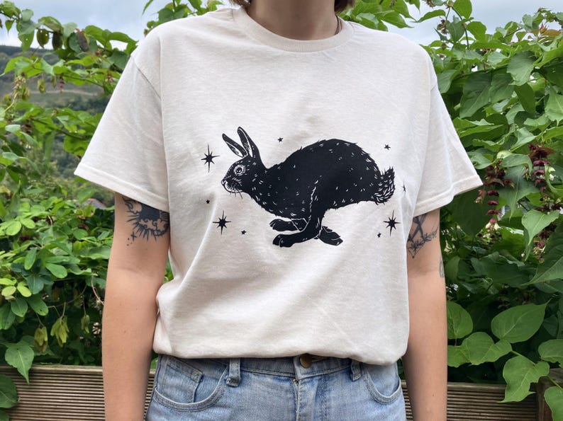 May include: A white t-shirt with a black silhouette of a rabbit running with stars printed on the front.