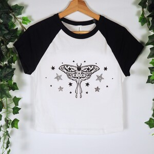 May include: White and black raglan style t-shirt with a black moth and star graphic. The moth has detailed wing patterns and is surrounded by stars and spiral designs. The shirt is hanging on a wooden hanger.