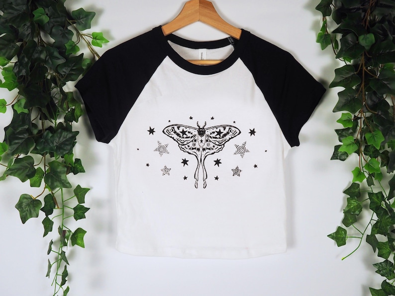 May include: White and black raglan style t-shirt with a black moth and star graphic. The moth has detailed wing patterns and is surrounded by stars and spiral designs. The shirt is hanging on a wooden hanger.
