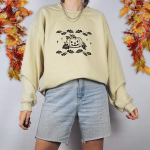 Pode incluir: Uma camisa de moletom bege claro com um gr&aacute;fico preto de uma ab&oacute;bora de Halloween com folhas e estrelas. A camisa de moletom est&aacute; sendo usada por uma pessoa que est&aacute; usando shorts jeans azul claro.