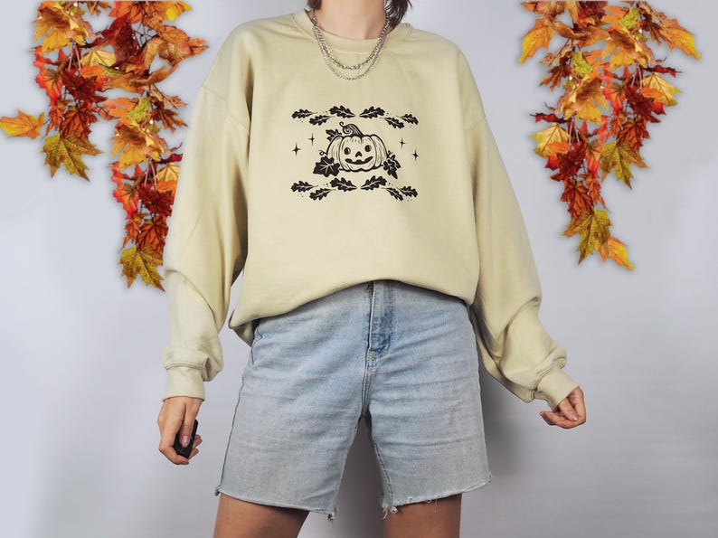 Pode incluir: Uma camisa de moletom bege claro com um gr&aacute;fico preto de uma ab&oacute;bora de Halloween com folhas e estrelas. A camisa de moletom est&aacute; sendo usada por uma pessoa que est&aacute; usando shorts jeans azul claro.