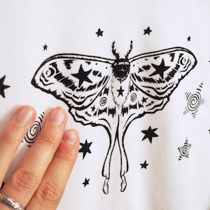 May include: White fabric featuring a black moth illustration with star and spiral accents. The moth has detailed wing patterns and star shapes. The design is surrounded by various sized stars and spiral designs.