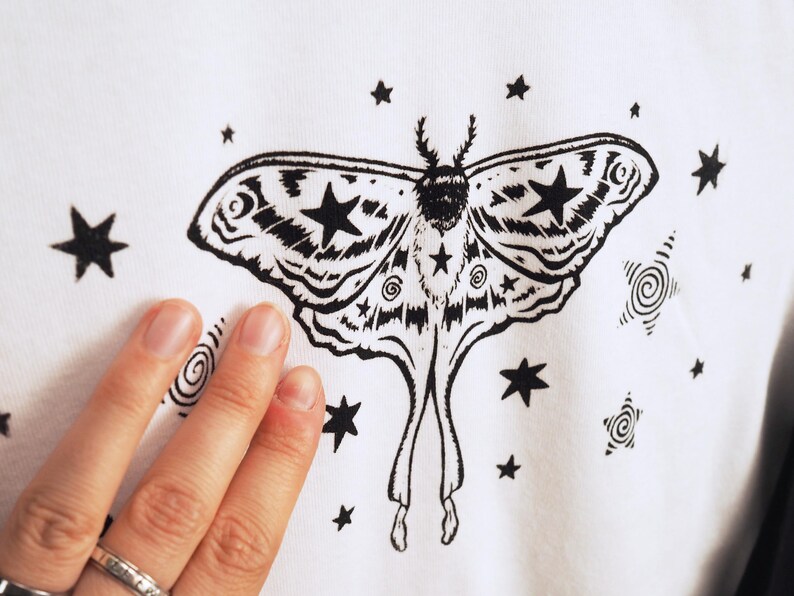 May include: White fabric featuring a black moth illustration with star and spiral accents. The moth has detailed wing patterns and star shapes. The design is surrounded by various sized stars and spiral designs.