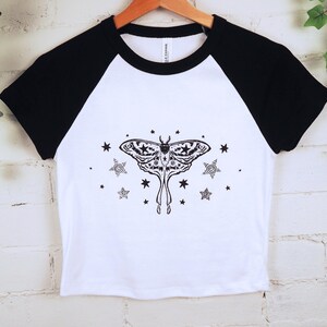 May include: White and black raglan crop top featuring a detailed black moth illustration with stars. The shirt has a crew neck and short sleeves. The design is centered on the front of the shirt.