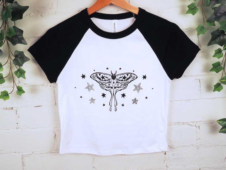 May include: White and black raglan crop top featuring a detailed black moth illustration with stars. The shirt has a crew neck and short sleeves. The design is centered on the front of the shirt.