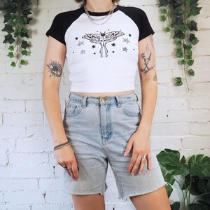 May include: White and black raglan crop top with a moth and star graphic. The top is paired with light wash denim shorts. The outfit is casual and suitable for everyday wear.