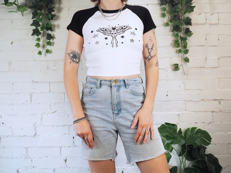 May include: White and black raglan crop top with a moth and star graphic. The top is paired with light wash denim shorts. The outfit is casual and suitable for everyday wear.