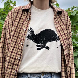 May include: A white t-shirt with a black silhouette of a rabbit running with stars printed on the front. The shirt is being worn over a brown and beige plaid shirt.
