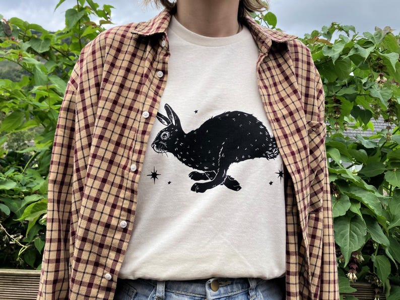 May include: A white t-shirt with a black silhouette of a rabbit running with stars printed on the front. The shirt is being worn over a brown and beige plaid shirt.