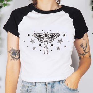 May include: White and black raglan style t-shirt featuring a detailed black moth illustration with star and spiral accents. The shirt has a round neckline and short sleeves. The design is centered on the front of the shirt.