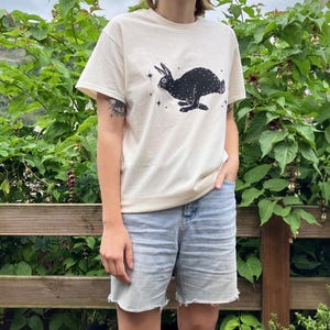 May include: A cream colored t-shirt with a black silhouette of a rabbit with stars printed on the front. The person wearing the shirt is standing in front of a green leafy bush.
