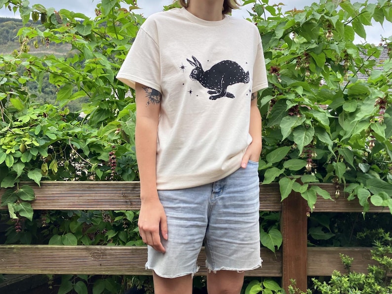 May include: A cream colored t-shirt with a black silhouette of a rabbit with stars printed on the front. The person wearing the shirt is standing in front of a green leafy bush.