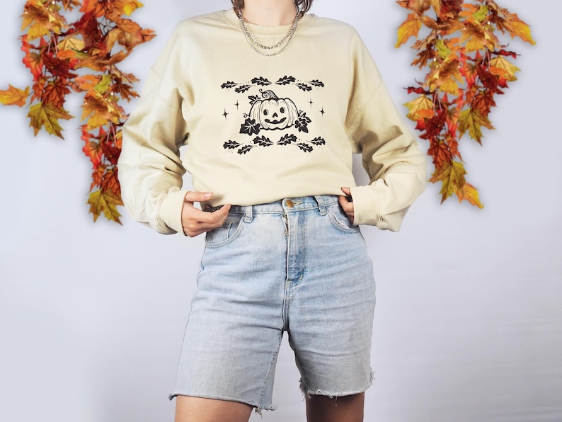 A woman wears a beige jumper with a pumpkin printed in black. The pumpkin has a face with a smile and is surrounded by  oak leaves.