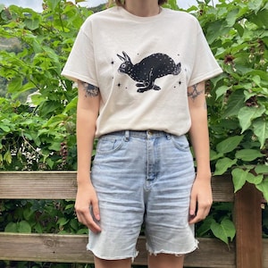 May include: A white t-shirt with a black rabbit silhouette and stars printed on the front. The rabbit is running and has long ears. The t-shirt is being worn by a person with denim shorts.