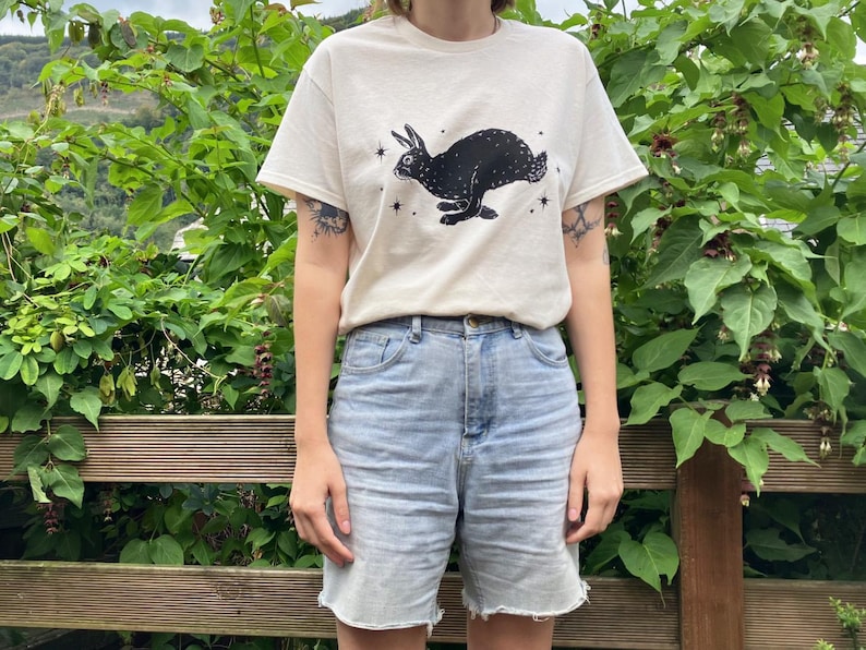 May include: A white t-shirt with a black rabbit silhouette and stars printed on the front. The rabbit is running and has long ears. The t-shirt is being worn by a person with denim shorts.