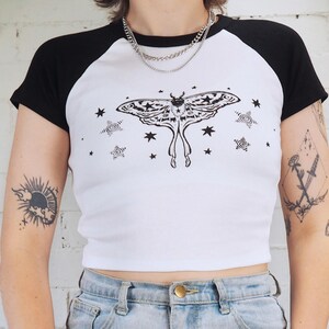 May include: White and black baseball style t-shirt with a large moth graphic on the front. The moth is black and white with star and swirl accents. The shirt has black sleeves and a white body.