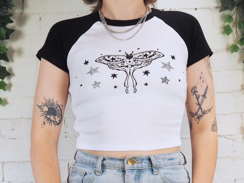 May include: White and black baseball style t-shirt with a large moth graphic on the front. The moth is black and white with star and swirl accents. The shirt has black sleeves and a white body.