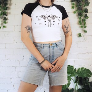 May include: White and black raglan crop top with a moth and star graphic. The shirt has black sleeves and a white body. The model is wearing light wash denim shorts. The background is a white brick wall with hanging greenery.