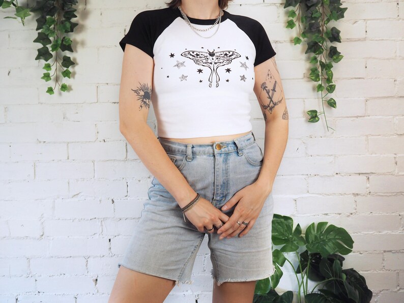 May include: White and black raglan crop top with a moth and star graphic. The shirt has black sleeves and a white body. The model is wearing light wash denim shorts. The background is a white brick wall with hanging greenery.