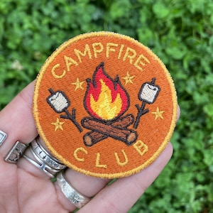 Può includere: Toppa rotonda ricamata arancione con le parole "CAMPFIRE CLUB" attorno a un disegno di falò. Il falò ha fiamme, tronchi, marshmallow su bastoncini e stelle. La toppa è tenuta in una mano.