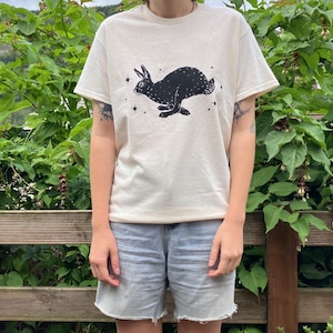 May include: A white t-shirt with a black rabbit illustration and stars printed on the front. The rabbit is running and has a starry tail.