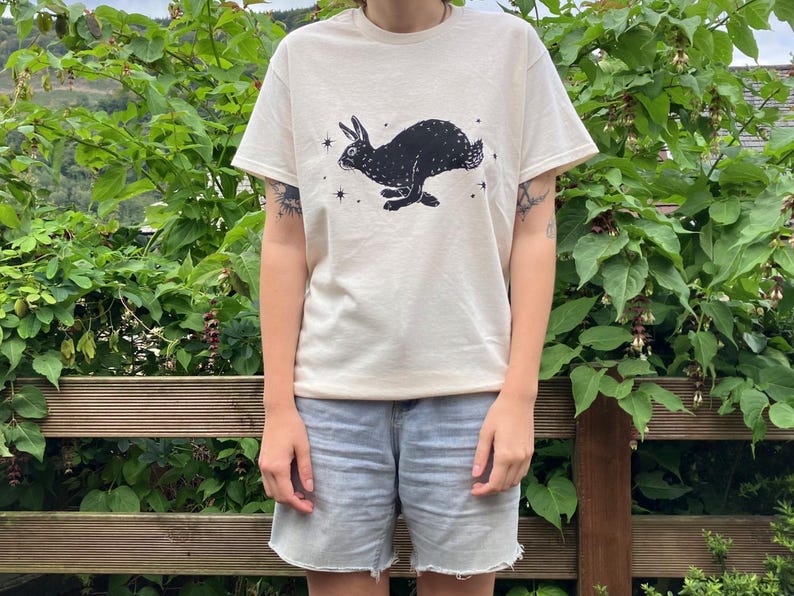 May include: A white t-shirt with a black rabbit illustration and stars printed on the front. The rabbit is running and has a starry tail.