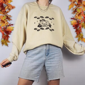 Pode incluir: Uma camisa de frio bege claro com um gr&aacute;fico preto e branco de uma ab&oacute;bora com folhas e estrelas. A camisa de frio est&aacute; sendo usada por uma pessoa que est&aacute; usando shorts jeans azul claro.