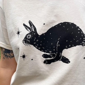 May include: A white t-shirt with a black silhouette of a rabbit running with stars printed on it.