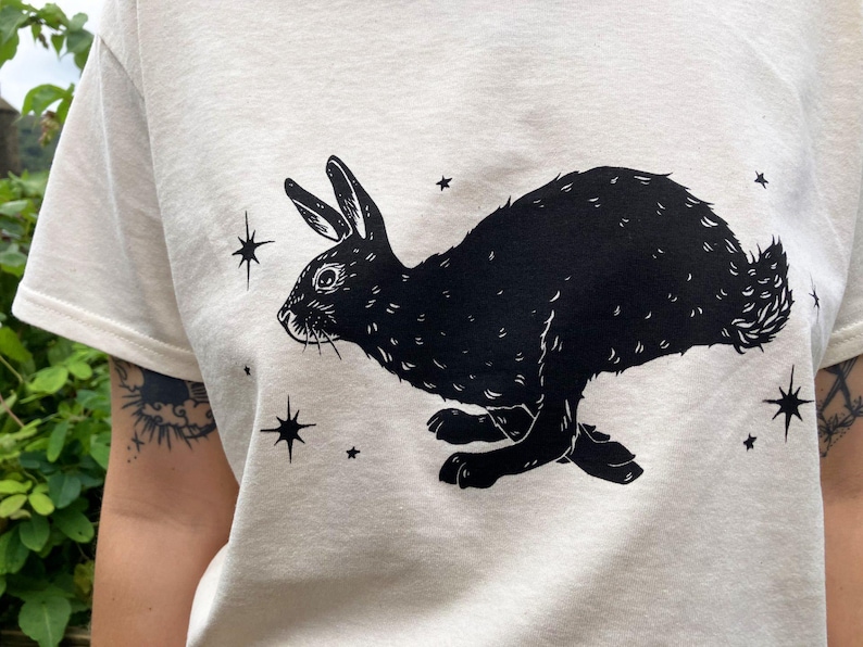 May include: A white t-shirt with a black silhouette of a rabbit running with stars printed on it.
