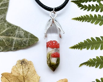 May include: A silver pendant necklace with a clear glass vial containing red and white mushrooms and green moss. The pendant is shaped like a teardrop and has a silver cap.