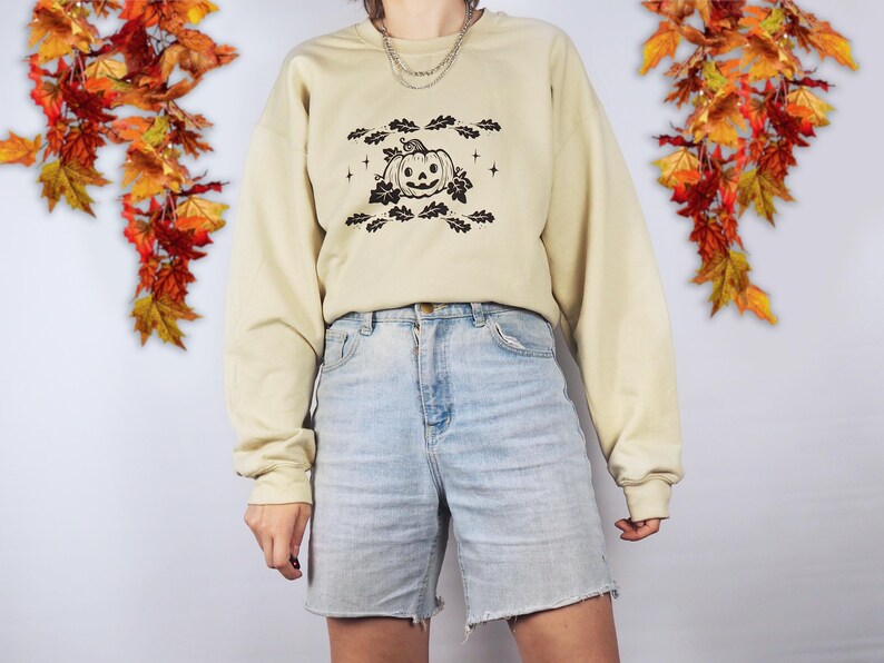 A photo of a woman wearing a beige jumper with an autumnal pumpkin surrounded by oak leaves printed on it.