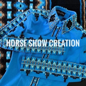 Op de afbeelding: Een blauw show shirt met zwarte en bruine bies en strass. Het shirt heeft een geometrisch patroon en de tekst "HORSE SHOW CREATION" op de voorkant.