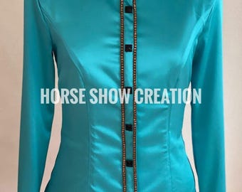 May include: A turquoise satin long-sleeve shirt with a black and white embellished collar and a beaded trim down the front. The shirt has a fitted silhouette and a button-up closure. The text "HORSE SHOW CREATION" is printed on the front of the shirt.