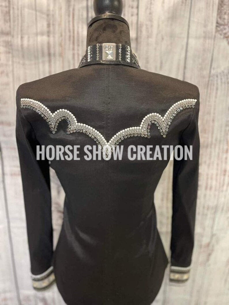 May include: Black show shirt with a white and silver trim and rhinestones. The shirt has a collar with a silver buckle and the text "HORSE SHOW CREATION" is printed on the back.
