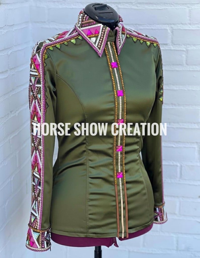 May include: Olive green long-sleeve shirt with decorative embellishments. The shirt features a pointed collar and cuffs with intricate beading and geometric patterns in pink, white, and gold. The text "HORSE SHOW CREATION" is displayed across the front.