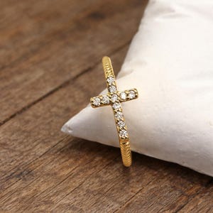 May include: A gold-colored ring with a cross design. The cross is adorned with small, clear gemstones. The ring is set on a white surface against a wooden background. The ring's band has a textured, ridged design.